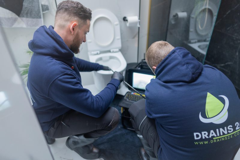 Drain engineer carrying out a CCTV drain survey at an outdoor inspection chamber using a drain camera and monitor unit beside a house