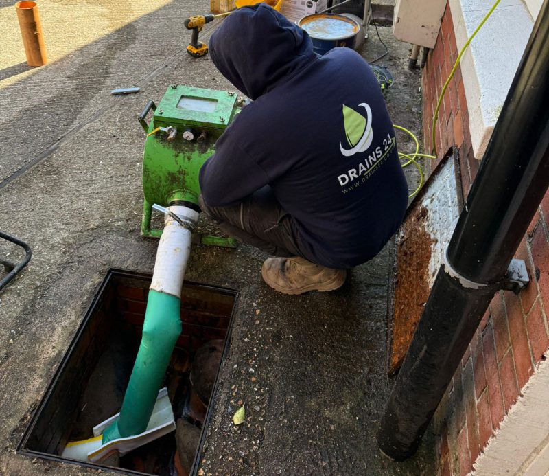 Drain engineer carrying out CIPP drain lining at an inspection chamber with liner material, reel and CCTV monitor beside a house